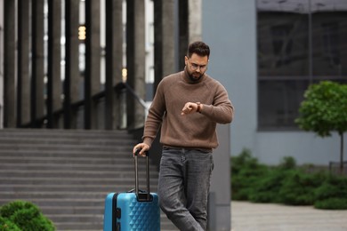 Photo of Being late. Worried man with suitcase looking at watch outdoors, space for text