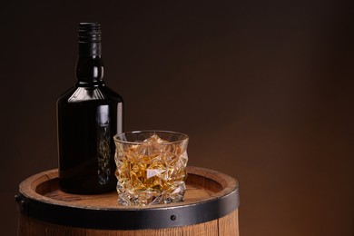 Photo of Whiskey with ice cubes in glass and bottle on wooden barrel against dark background, space for text