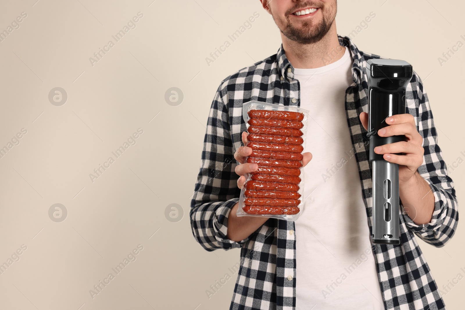 Photo of Smiling man holding sous vide cooker and sausages in vacuum pack on beige background, closeup. Space for text