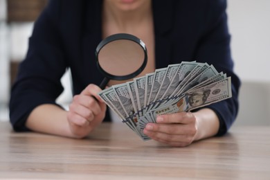 Photo of Expert authenticating 100 dollar banknotes with magnifying glass at table in office, closeup. Fake money concept