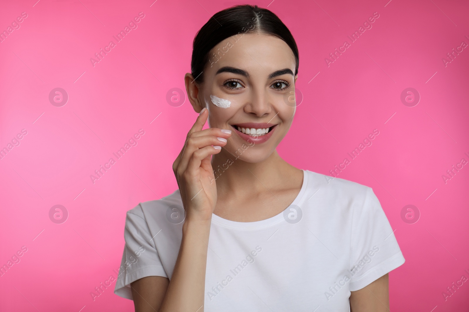 Photo of Young woman applying facial cream on pink background