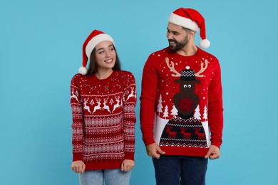 Photo of Happy young couple in Santa hats showing Christmas sweaters on light blue background