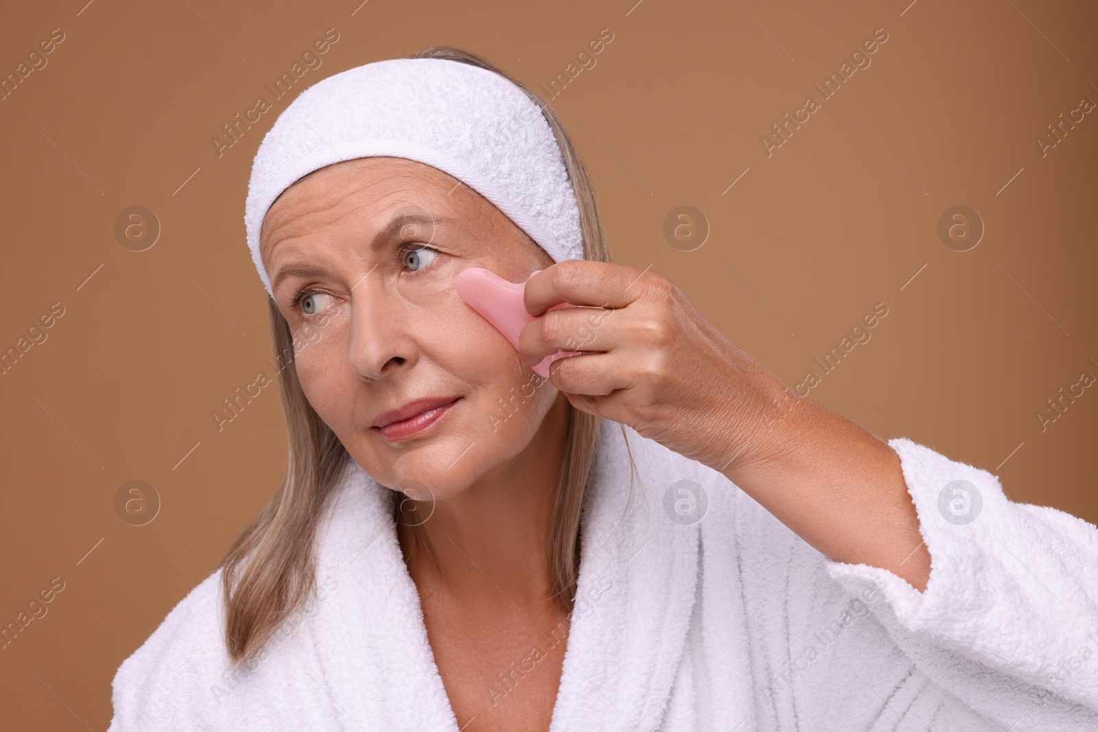 Photo of Woman massaging her face with rose quartz gua sha tool on brown background