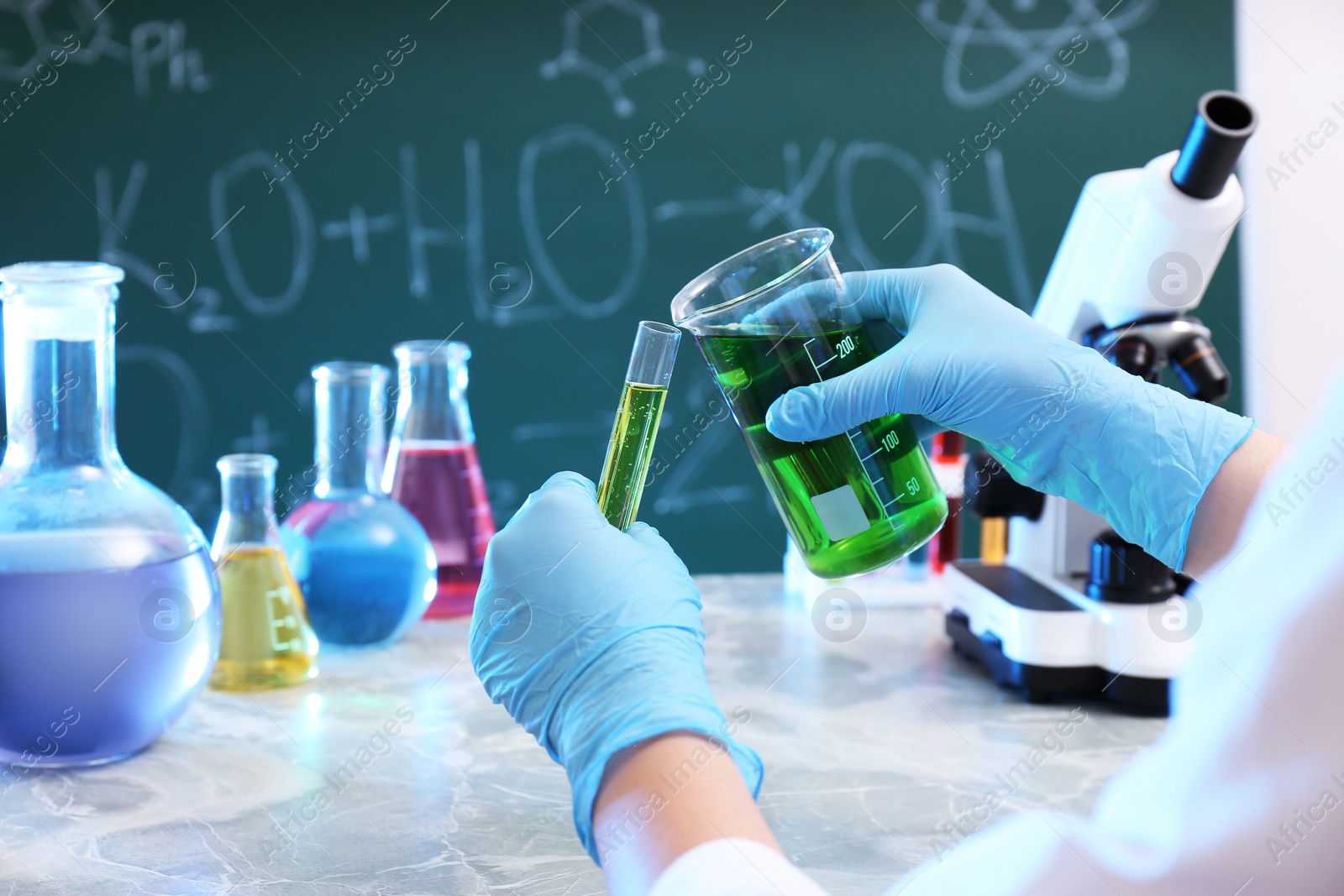 Photo of Scientist pouring liquid into test tube at table against chalkboard, closeup. Chemistry glassware