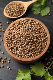 Photo of Dried coriander seeds in bowl, spoon and green leaves on dark gray textured table, above view