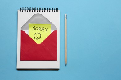 Image of Note with word Sorry and drawn sad face in envelope, notebook and pencil on light blue background, flat lay. Space for text