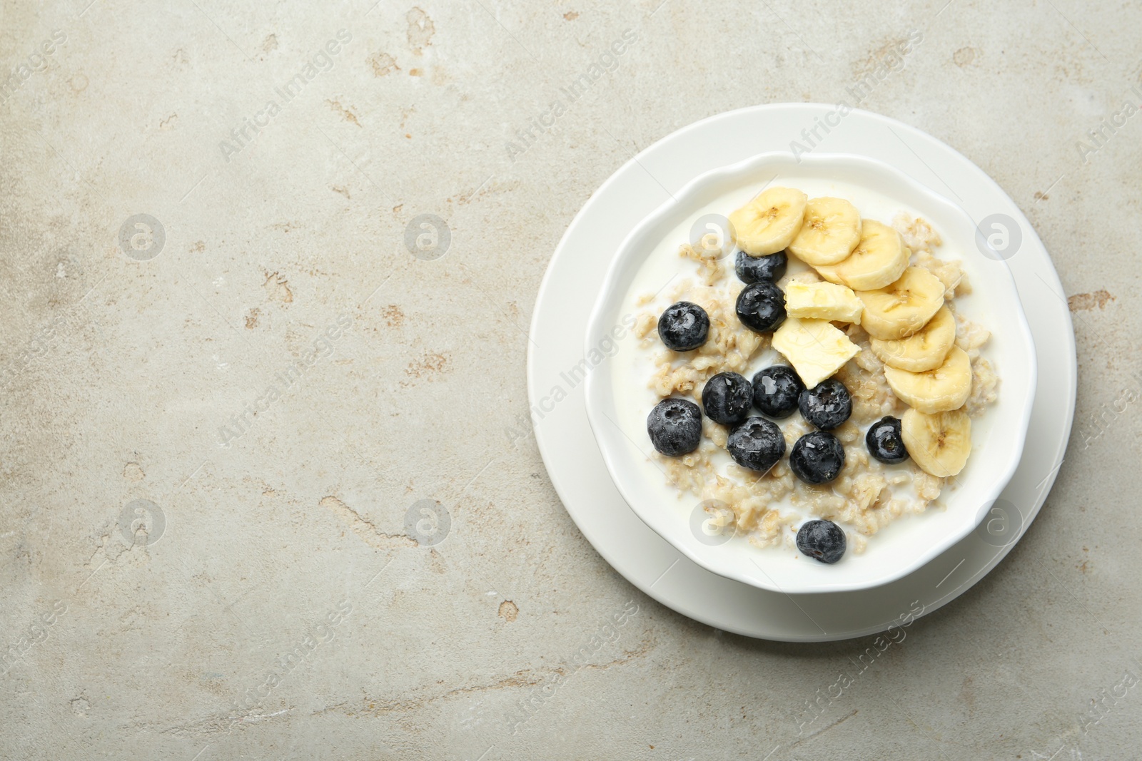 Photo of Tasty oatmeal with banana, blueberries, butter and milk served in bowl on light grey table, top view. Space for text