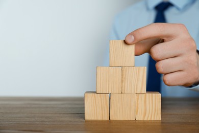 Photo of Businessman building pyramid of blank cubes on wooden table against light background, closeup. Space for text