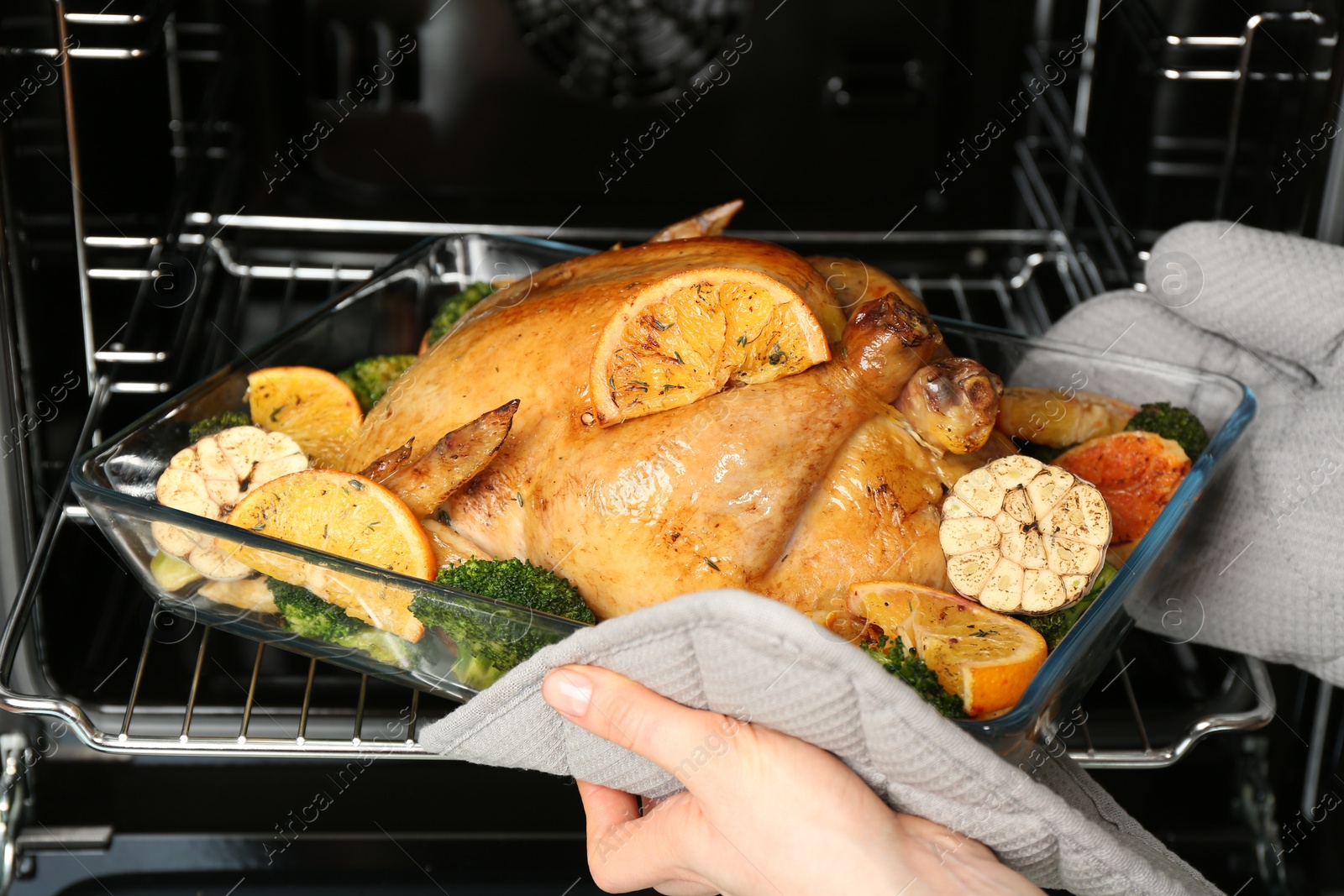 Photo of Woman taking baked chicken with oranges and vegetables out of oven, closeup