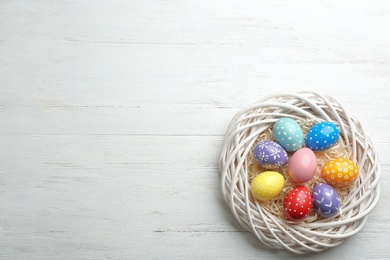 Wicker nest with painted Easter eggs on wooden table, top view. Space for text