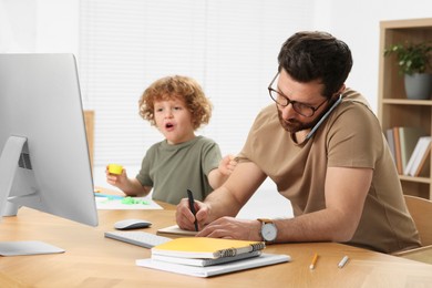 Photo of Little boy bothering his father at home. Man working remotely at desk