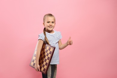 Cute girl with chessboard showing thumb up on pink background