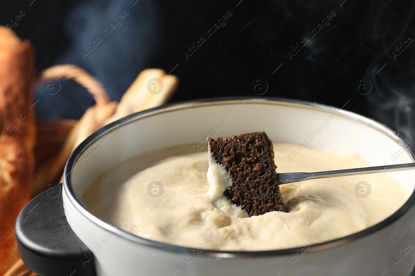 Photo of Dipping piece of bread into fondue pot with melted cheese on black background, closeup