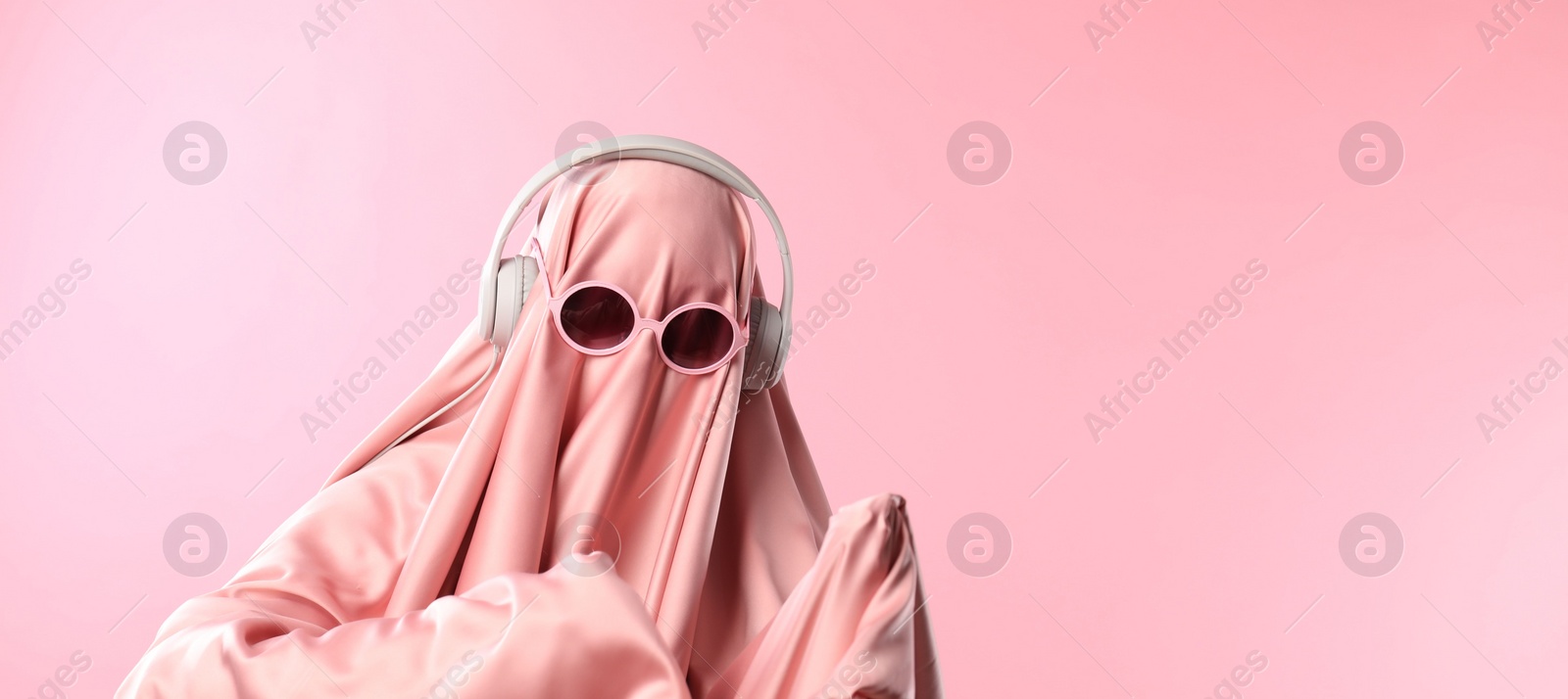 Photo of Glamorous ghost. Woman in sheet with sunglasses and headphones on pink background, space for text