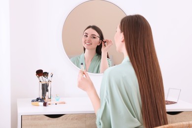 Photo of Beautiful young woman applying eyebrow gel at dressing table indoors