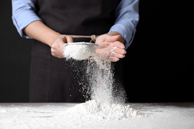 Photo of Woman sifting wheat flour at table against black background, closeup