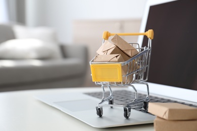 Photo of Internet shopping. Laptop and small cart with boxes on table indoors, space for text