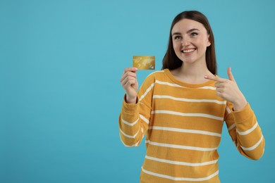 Happy woman pointing at credit card on light blue background, space for text