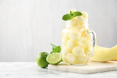 Photo of Mason jar of melon ball cocktail served with mint and lime on white marble table. Space for text