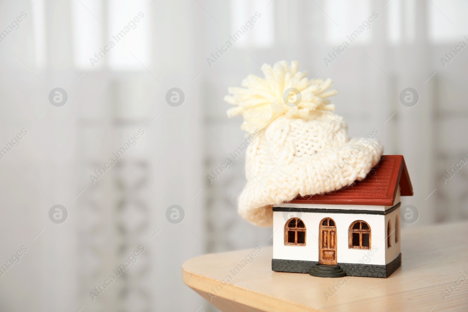 Photo of House model with knitted hat on table against blurred background, space for text. Heating concept