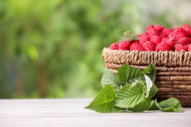 Wicker basket with tasty ripe raspberries and leaves on white wooden table against blurred green background, space for text