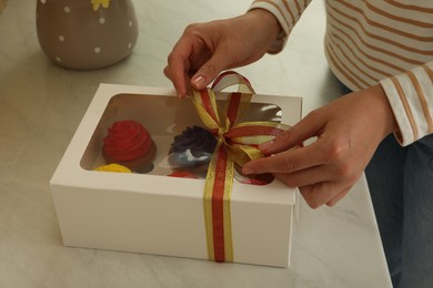 Photo of Woman tying bow on box with delicious colorful cupcakes at white table, closeup