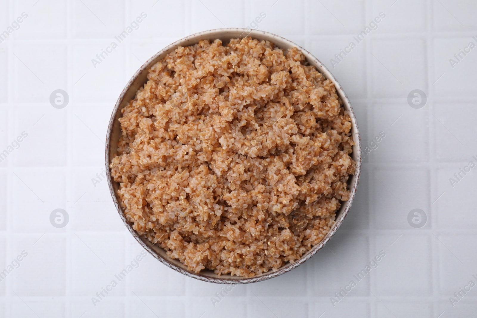 Photo of Tasty wheat porridge in bowl on white tiled table, top view