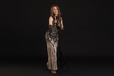 Beautiful young woman in stylish dress with microphone singing on black background