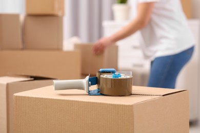 Photo of Woman sorting boxes indoors, focus on dispenser with roll of adhesive tape