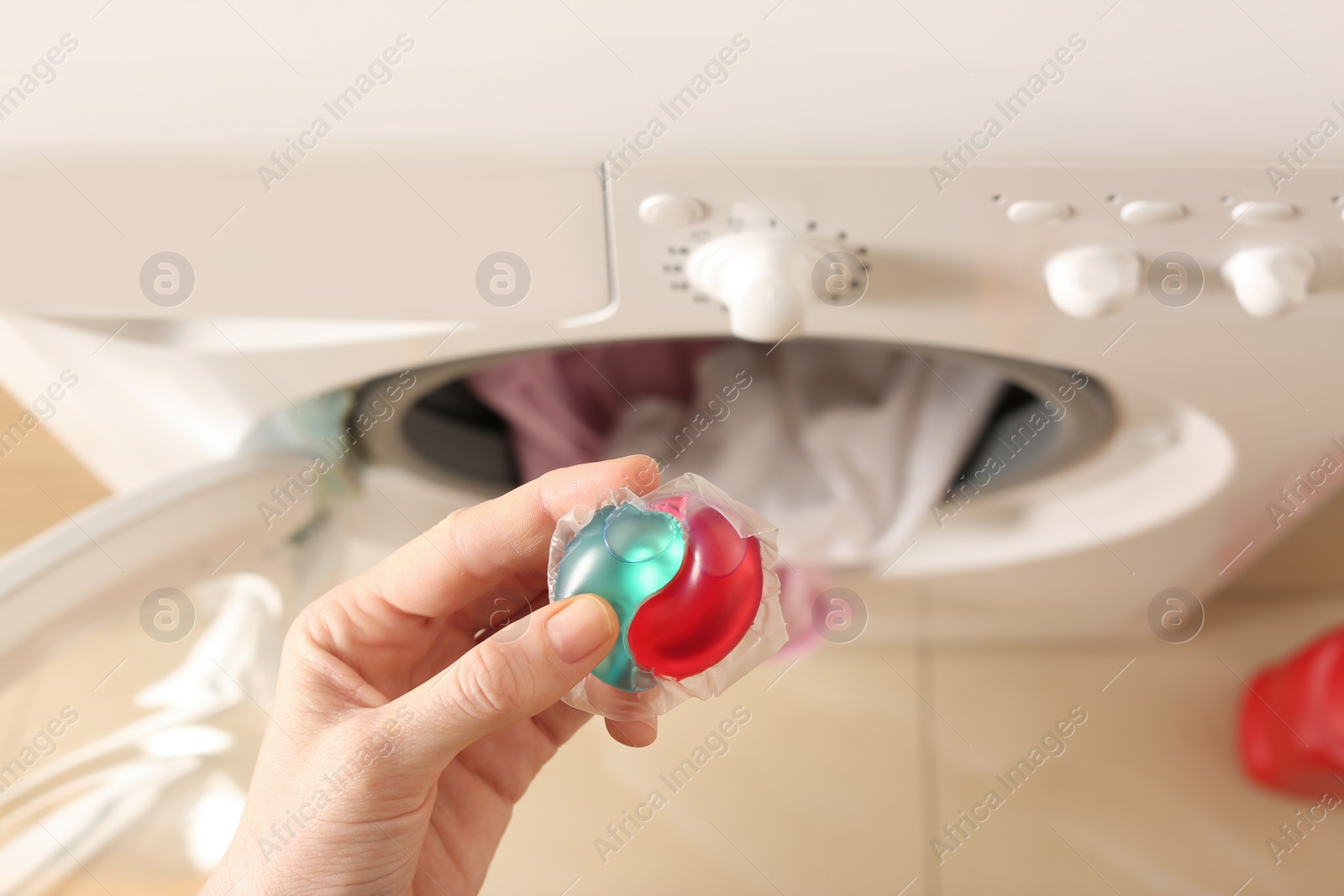 Photo of Woman holding laundry detergent capsule near washing machine indoors, top view