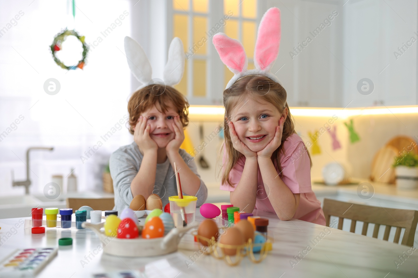 Photo of Painting Easter eggs. Cute children with bunny ears at white marble table in kitchen