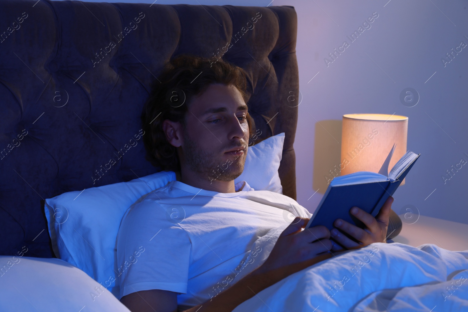 Photo of Handsome young man reading book in dark room at night. Bedtime