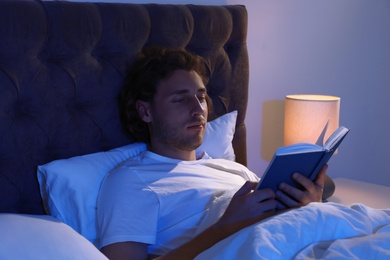 Photo of Handsome young man reading book in dark room at night. Bedtime