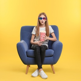 Photo of Emotional teenage girl with 3D glasses and popcorn sitting in armchair during cinema show on color background