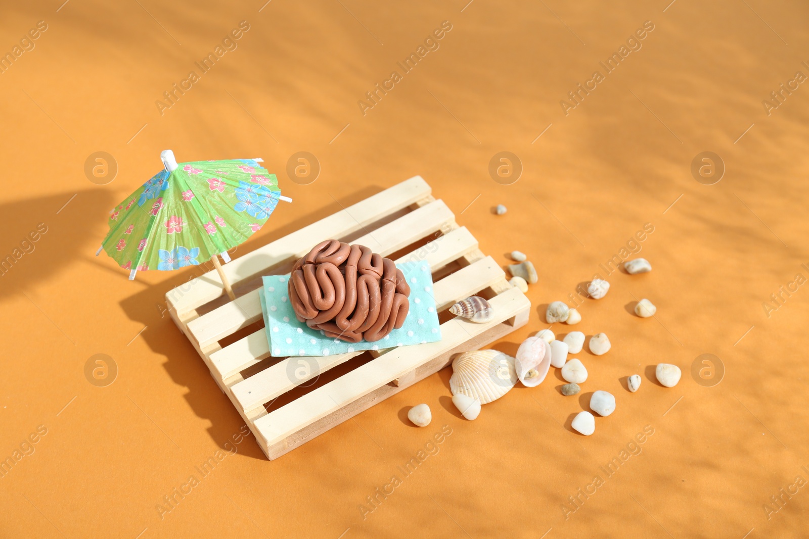 Photo of Brain made of plasticine on mini wooden sunbed under umbrella against orange background