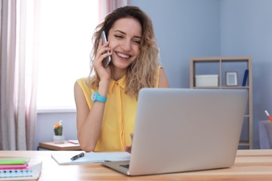 Young woman talking on mobile phone while working with laptop at desk. Home office