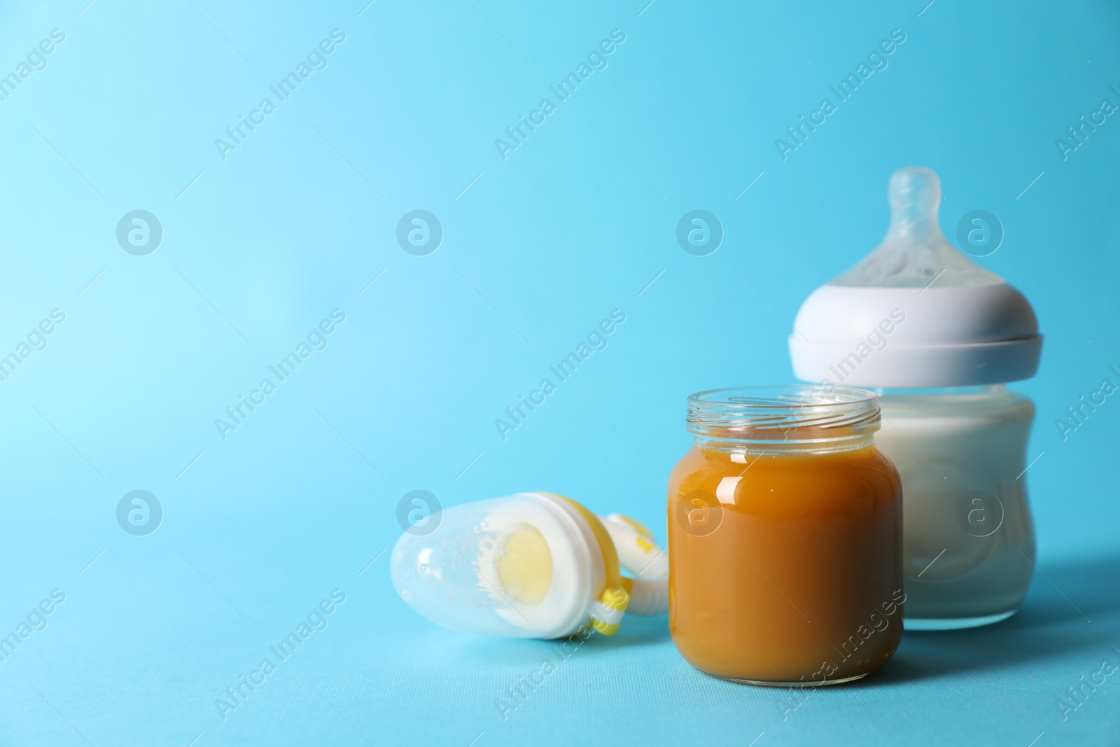 Photo of Jar with healthy baby food, bottle of milk and fruit feeder on light blue background. Space for text