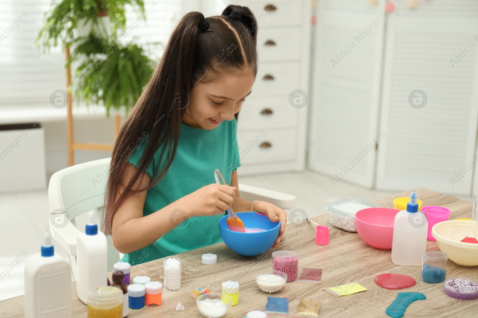 Photo of Cute little girl mixing ingredients with silicone spatula at table in room. DIY slime toy