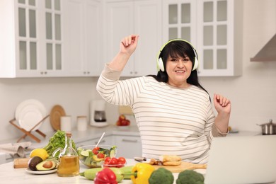 Photo of Happy overweight woman with headphones dancing while cooking in kitchen