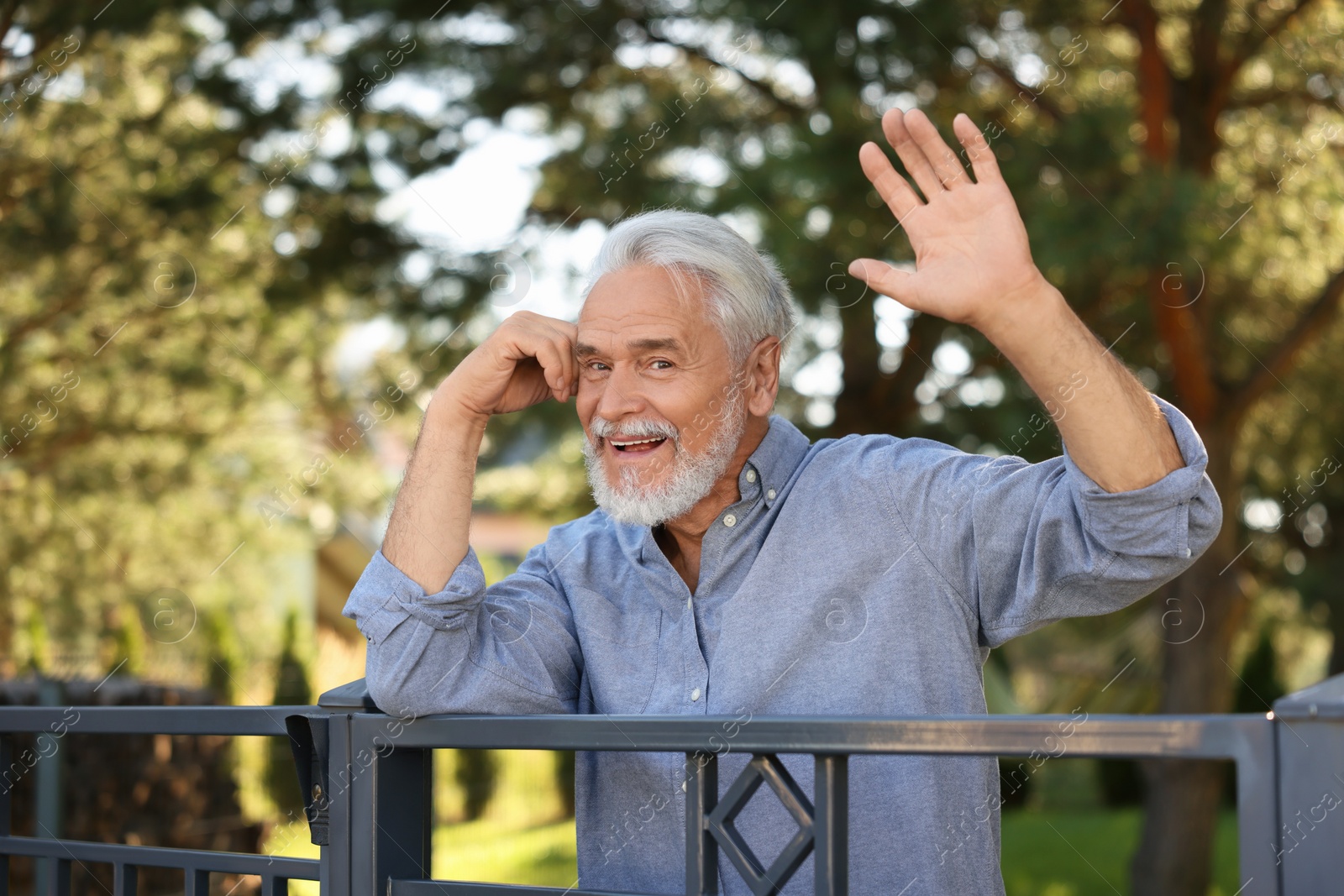 Photo of Neighbor greeting. Happy senior man waving near fence outdoors