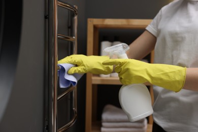 Photo of Woman cleaning heated towel rail with sprayer and rag indoors, closeup