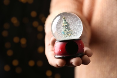 Photo of Woman holding snow globe with Christmas tree against festive lights, closeup