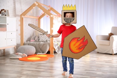 Photo of Cute little boy playing with cardboard armor in bedroom