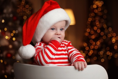 Cute little baby in Santa hat on chair against blurred festive lights. Christmas celebration