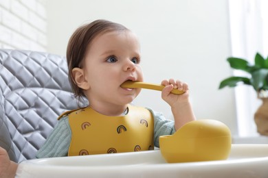 Photo of Cute little baby nibbling spoon in high chair indoors.