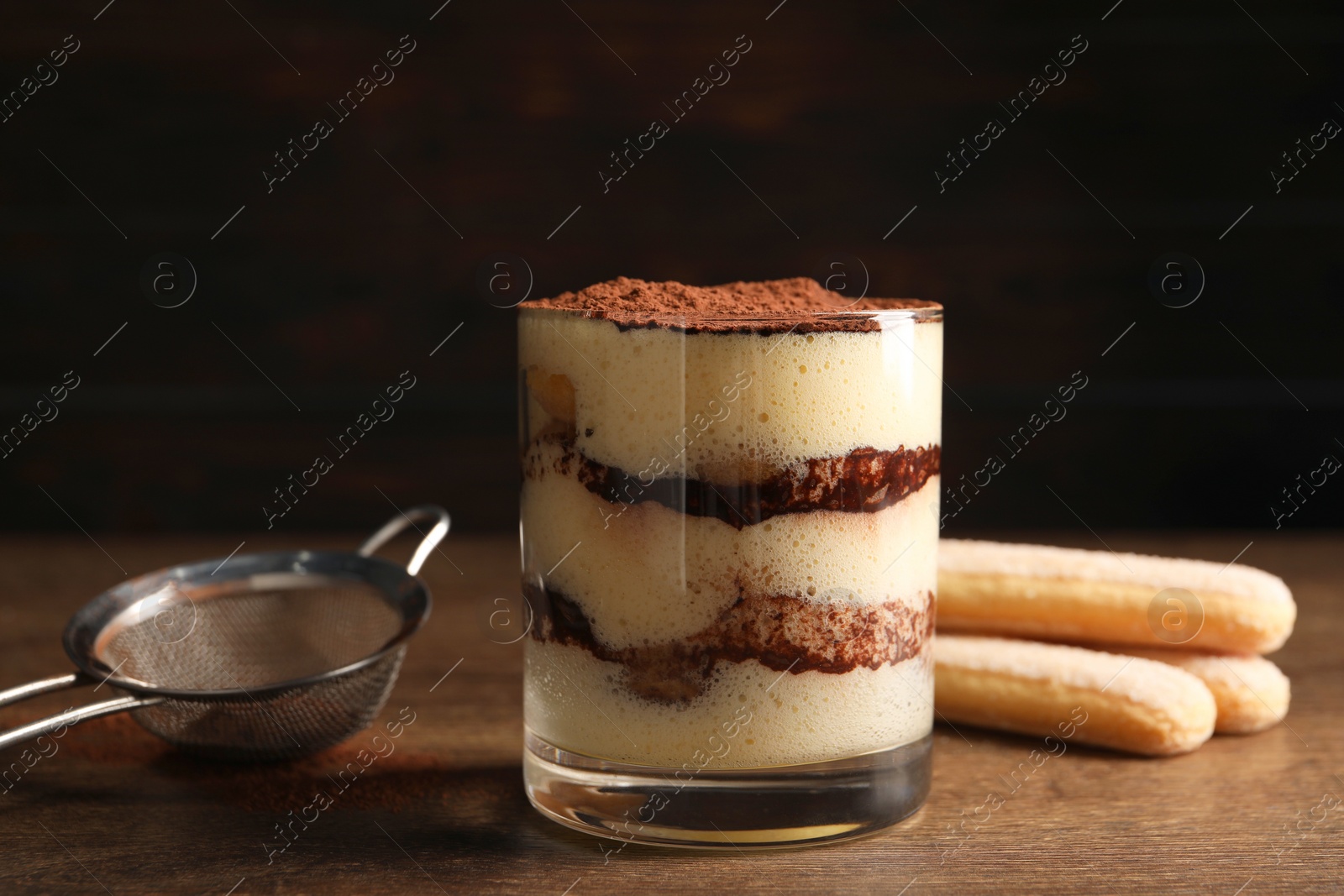 Photo of Delicious tiramisu in glass, biscuits and sieve on wooden table, closeup