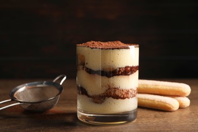 Photo of Delicious tiramisu in glass, biscuits and sieve on wooden table, closeup