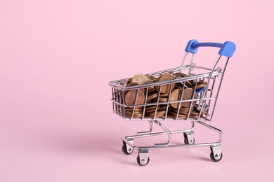 Photo of Small metal shopping cart with coins on pink background, space for text