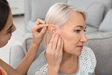 Young woman putting hearing aid in mother's ear indoors