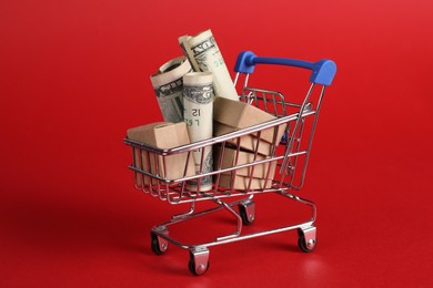 Photo of Small metal shopping cart with cardboard boxes and dollar banknotes on red background